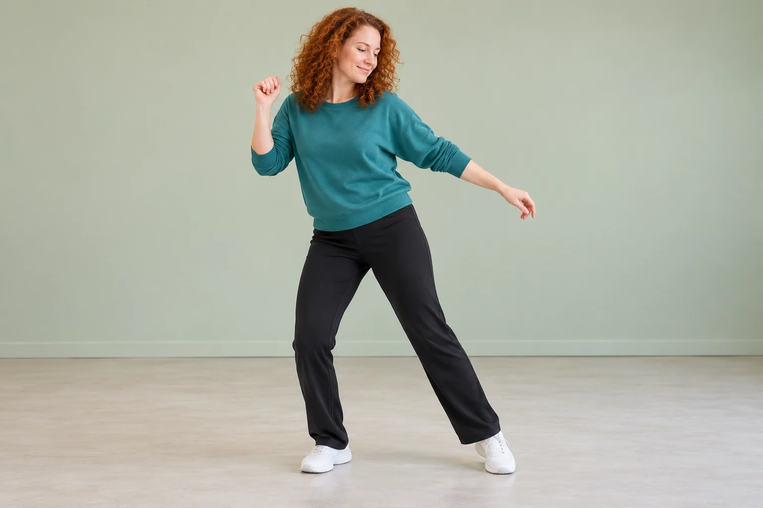 Auburn-haired woman in a teal sweatshirt dancing against a pale sage studio wall